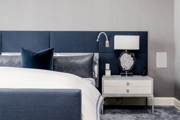 A modern bedroom features a blue upholstered bed, navy and gray pillows, a nightstand with a lamp, a framed art piece, and a mounted reading light.