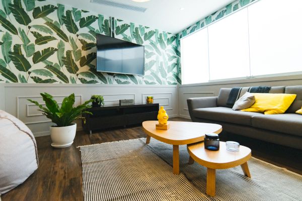 A stylish living room with tropical leaf wallpaper, gray sofa, yellow pillows, wooden coffee tables, potted plants, and a wall-mounted TV.