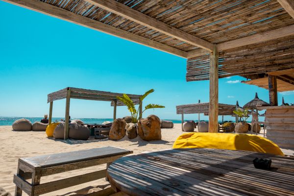 A sunny beach scene with shaded wooden structures, yellow and brown bean bags, tables, and clear blue sky.