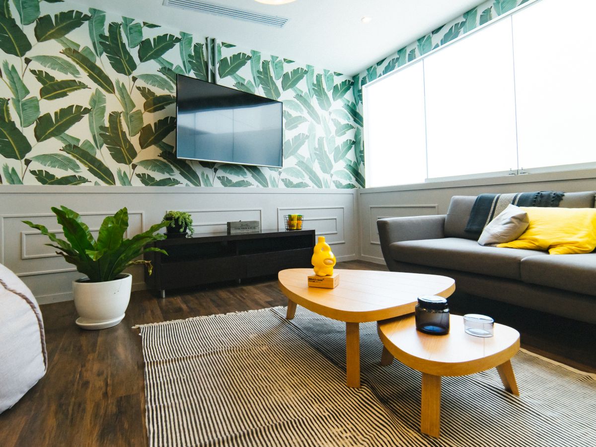 A modern living room with leaf-patterned wallpaper, a wall-mounted TV, a grey sofa with yellow cushions, a coffee table, and indoor plants.