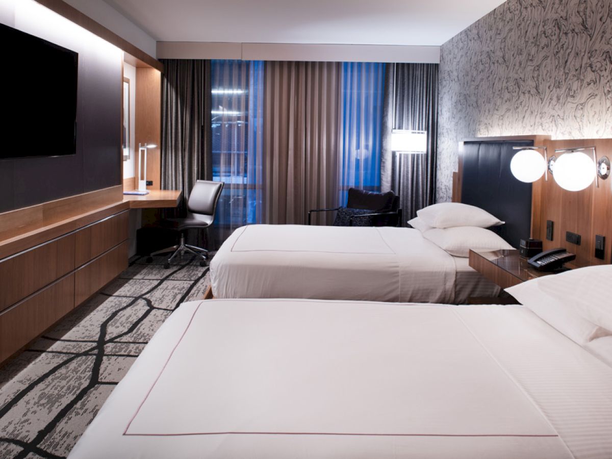A modern hotel room featuring two double beds, a large wall-mounted TV, a desk with a chair, ambient lighting, and patterned decor.