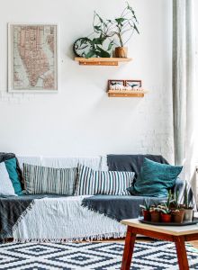 A cozy living room with a couch, pillows, plants, a geometric rug, wall art, and large windows allowing natural light to flood the space.