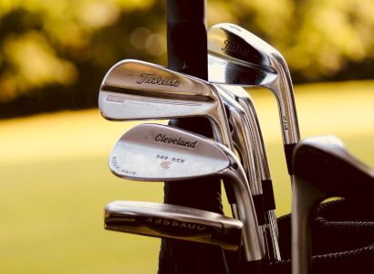 The image shows a close-up of golf clubs, including brands like Titleist and Cleveland, in a golf bag with a blurred green background.