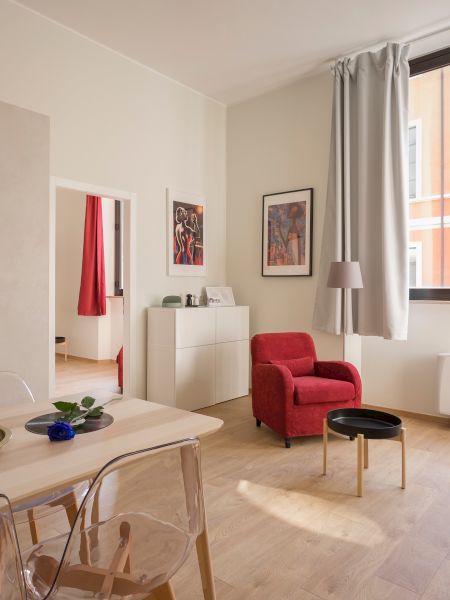 A bright living room with a red armchair, patterned sofa, dining table, and kitchen area. There is a large window and art on the walls.