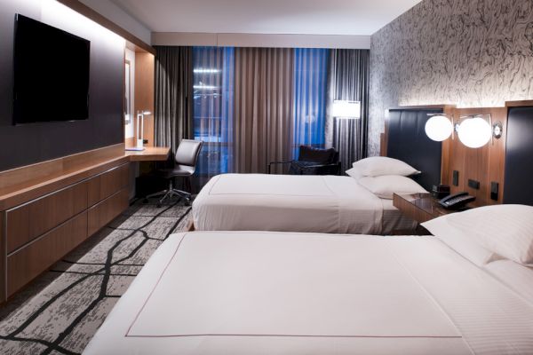 A modern hotel room featuring two beds, a large TV, a desk by the window, and ambient lighting; the decor is sleek and contemporary.