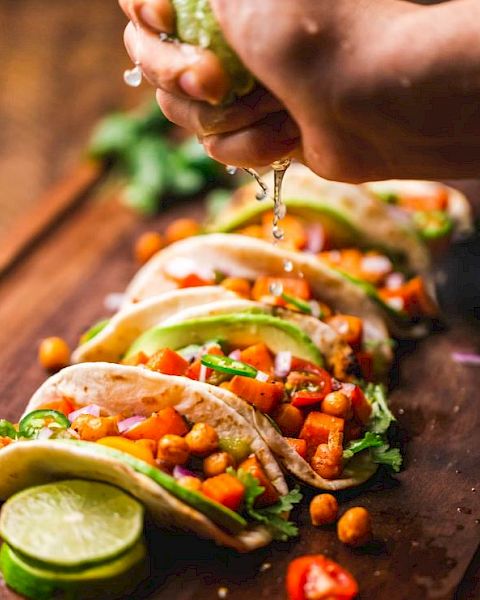 The image shows a person squeezing lime juice over a row of vegetable-filled tacos on a wooden surface with garnish, ready to be served.