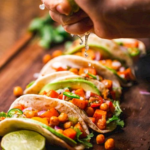 The image shows a person squeezing lime juice over a row of vegetable-filled tacos on a wooden surface with garnish, ready to be served.