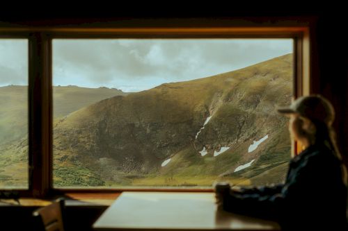 A person sits at a table, gazing out a large window at a mountainous landscape with patches of snow.