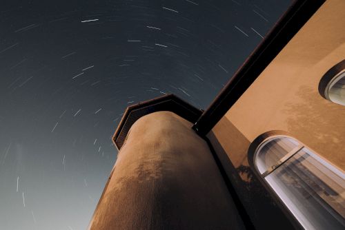 The image shows a tall building with a curved structure and a window, set against a night sky with star trails indicating long exposure photography.