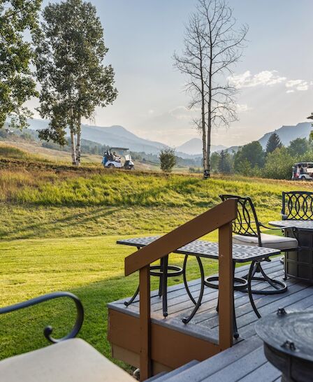 A peaceful backyard deck overlooking a grassy field and distant mountains, with patio furniture and a couple of glasses on the table.