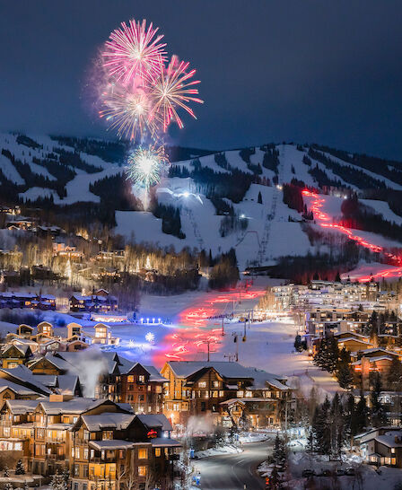 A winter landscape shows a mountain resort with fireworks overhead, illuminated buildings, and ski trails highlighted by lights.
