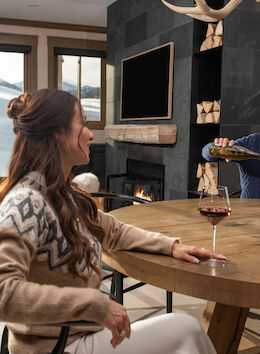 A cozy indoor setting shows a woman enjoying wine while a man pours another glass. Snowy mountains are visible through the window.
