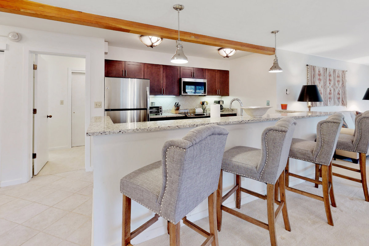 This image shows a modern kitchen with a breakfast bar, four high-backed chairs, stainless steel appliances, and pendant lighting.