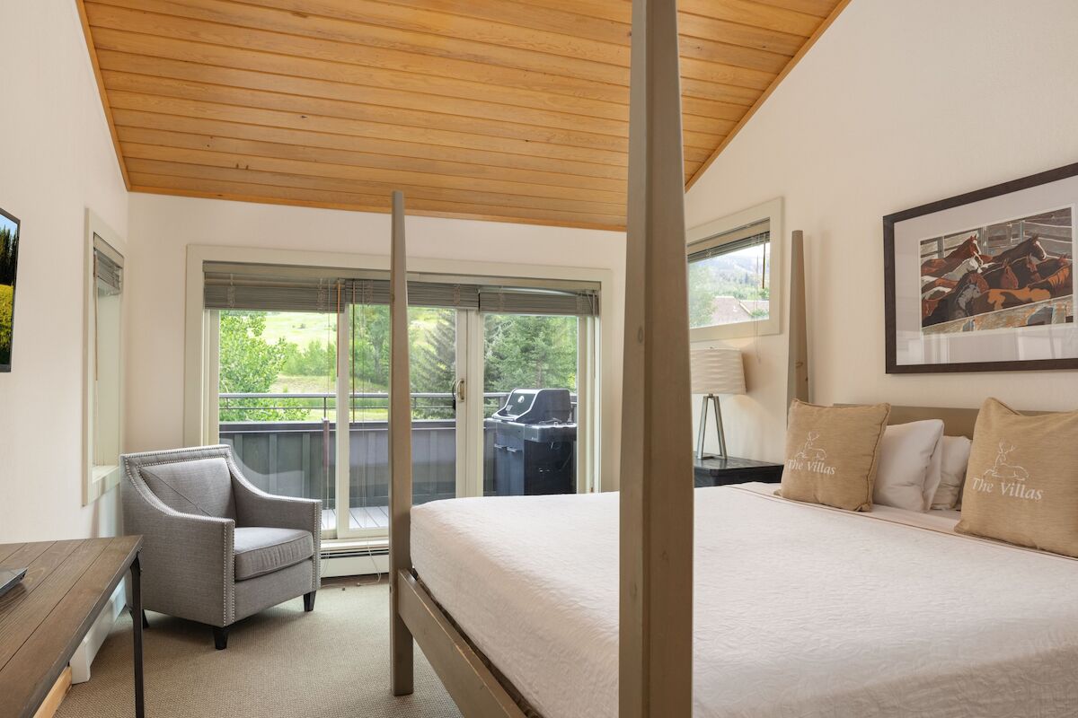 The image shows a cozy bedroom with a wooden ceiling, a queen bed, a gray chair, and large windows offering greenery views.