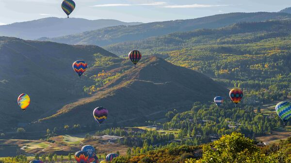 Hot air balloons float over a hilly landscape with trees, valleys, and a clear sky.