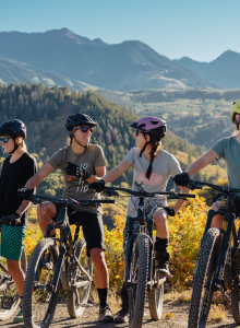 A group of six people are dressed in biking gear, standing next to their mountain bikes with scenic mountains in the background, chatting and smiling.
