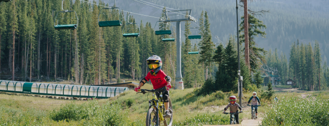 Three people are mountain biking on a trail with wooden bridges in a forested area, with ski lifts and distant hills in the background.