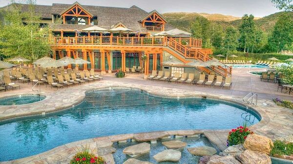 The image shows a luxurious outdoor pool area in front of a large building with wooden architecture, surrounded by hills and greenery, ending the sentence.