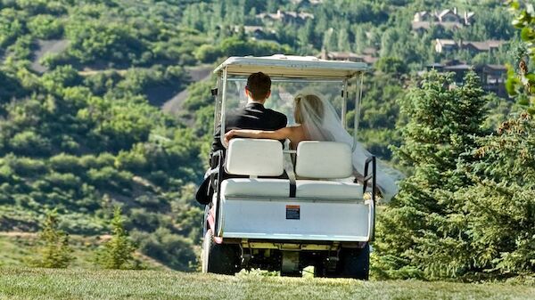 A couple, possibly newlyweds, are riding in a golf cart through a scenic landscape with lush greenery and distant houses.