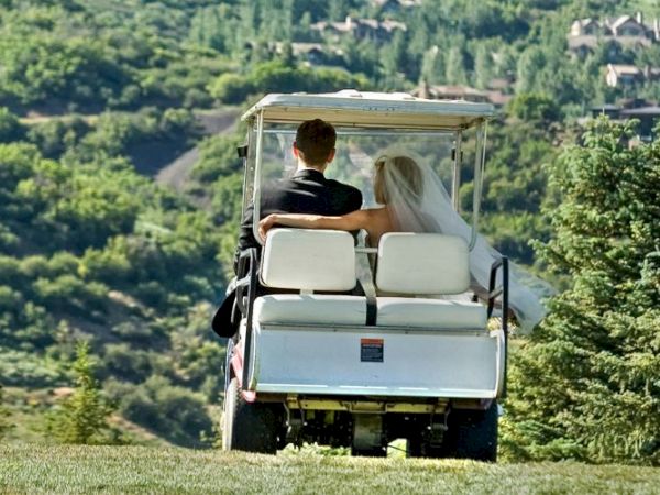 A couple, possibly newlyweds, are riding in a golf cart through a scenic landscape with lush greenery and distant houses.