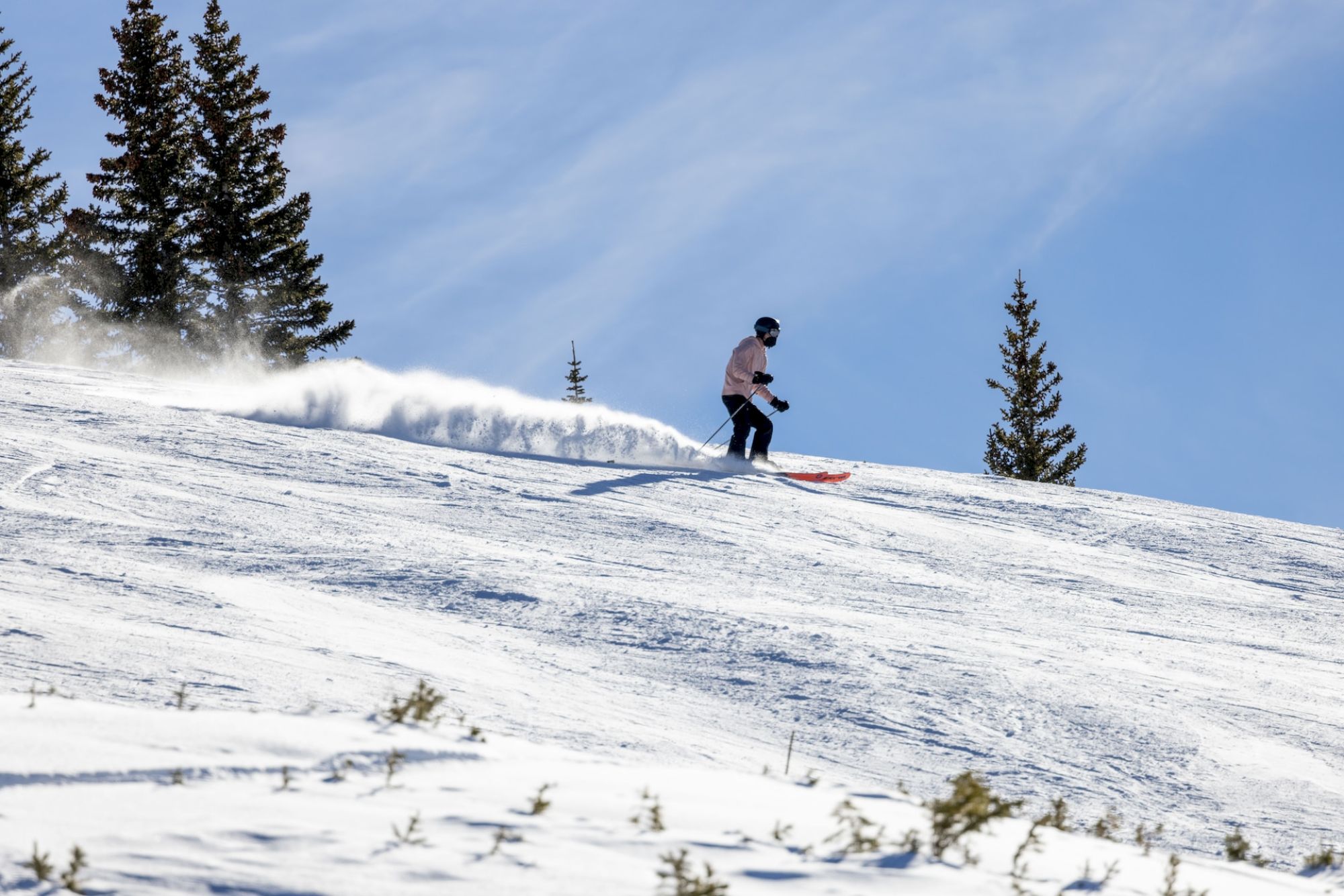 A skier is gliding down a snowy slope surrounded by trees, under a clear blue sky. Snow is being kicked up behind the skier as he moves.
