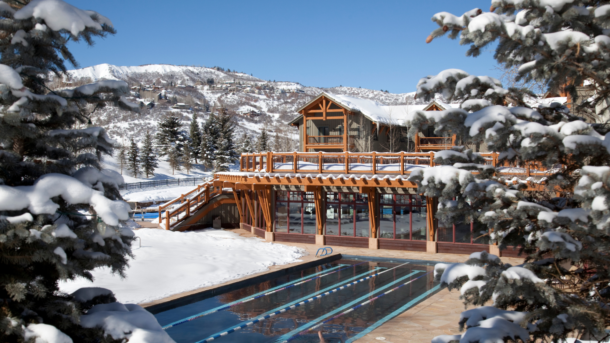 Snow-covered landscape with a wooden lodge, outdoor swimming pool, trees, and snowy mountains in the background, bathed in bright sunlight.