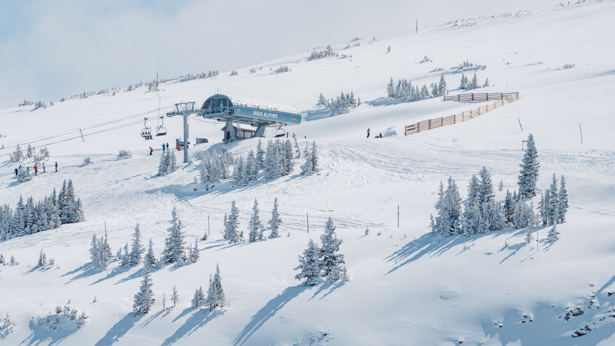 A snowy ski resort with a ski lift, skiers, and snow-covered trees on a mountainous landscape under a clear sky.