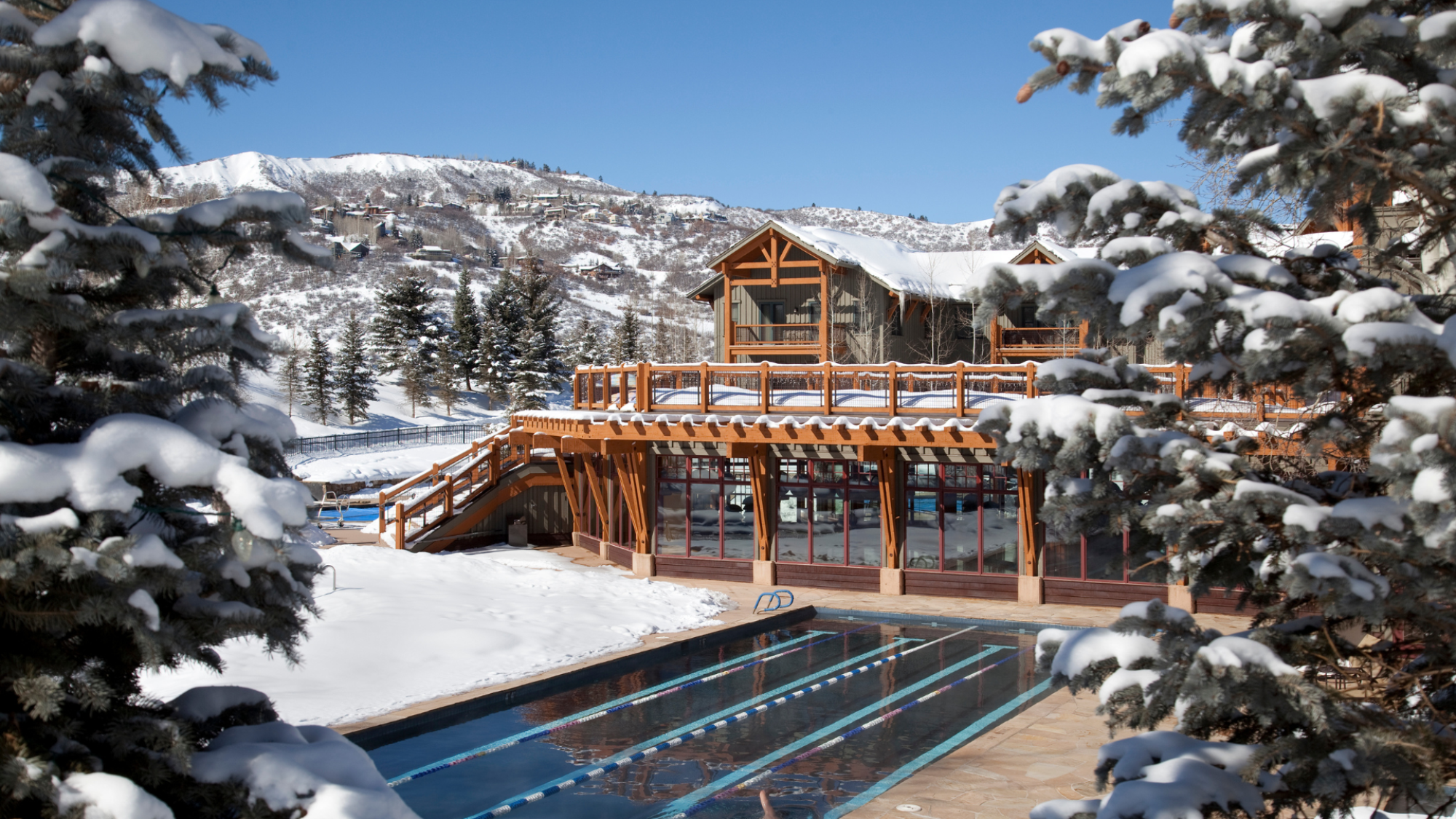 A snowy mountain scene featuring a wooden lodge and a swimming pool, surrounded by trees covered in snow, with clear blue skies overhead.