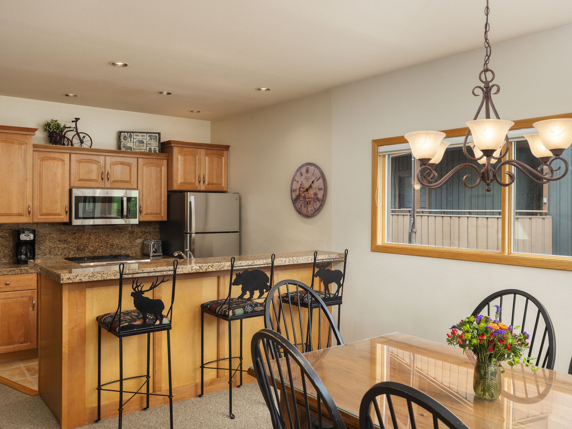 A cozy kitchen with wooden cabinets, bar stools, a dining table with flowers, and a chandelier, creating a warm atmosphere.