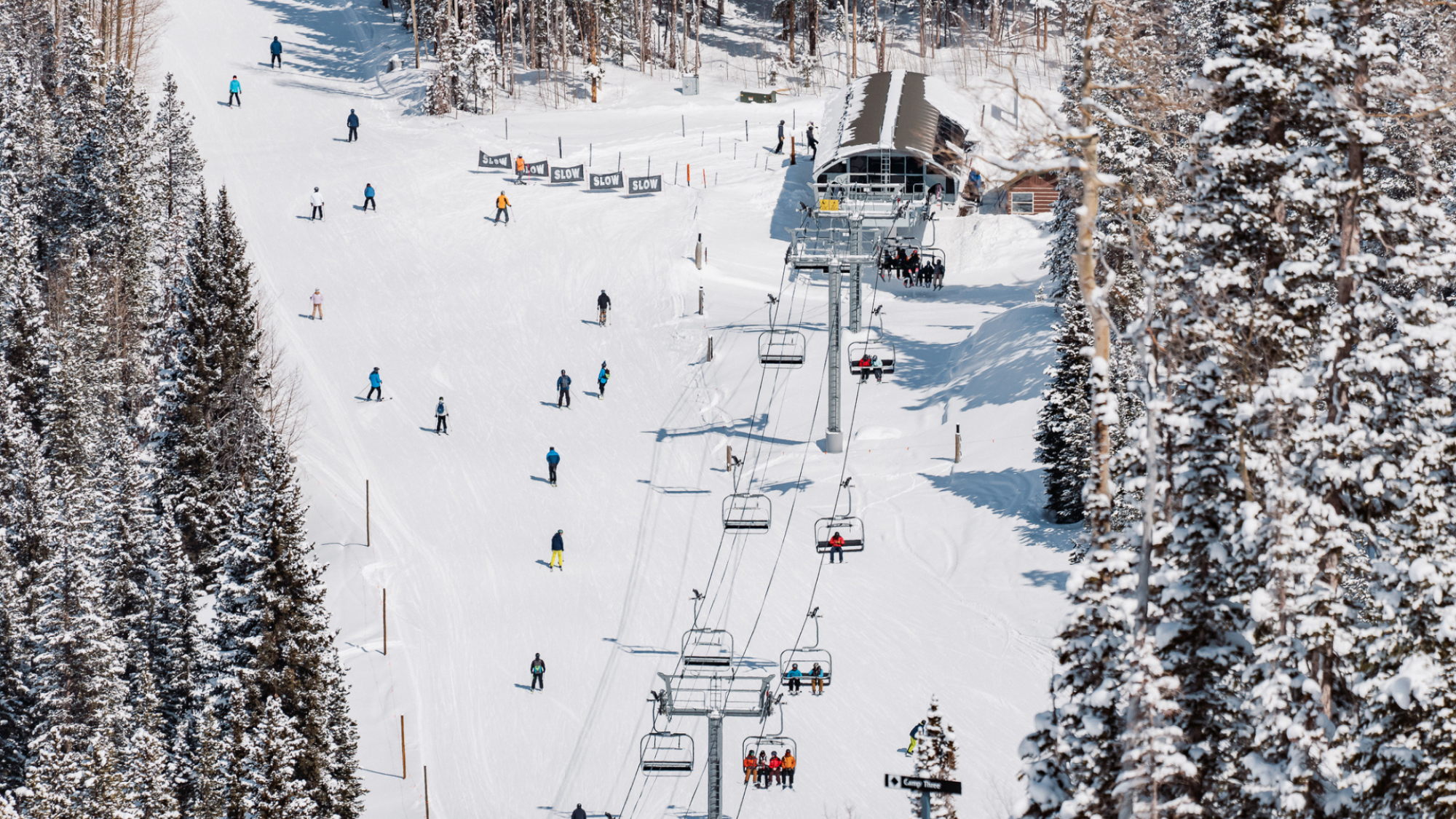 A snowy ski resort with skiers and snowboarders on slopes, surrounded by snow-covered trees and a ski lift leading to a building.