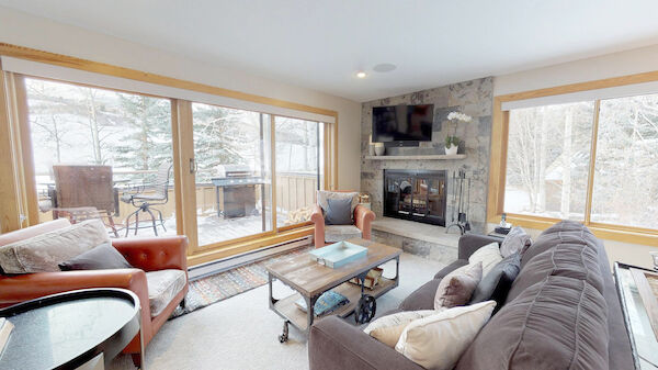 A cozy living room with large windows, a fireplace, a TV above the mantle, a gray couch, armchairs, and a coffee table, overlooking a snowy patio.