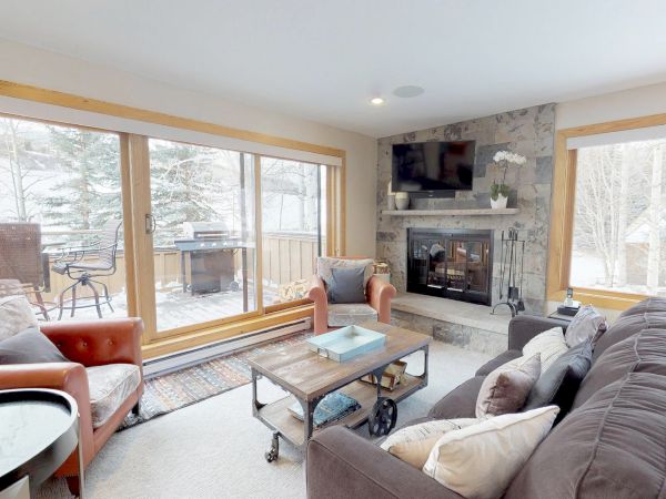A cozy living room with large windows, a fireplace, a TV above the mantle, a gray couch, armchairs, and a coffee table, overlooking a snowy patio.