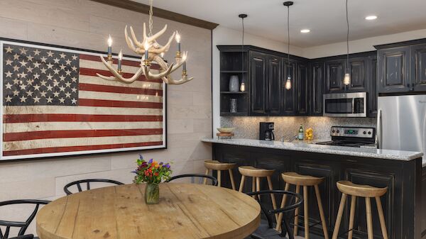 A rustic kitchen with a large American flag on the wall, wooden table, antler chandelier, black cabinets, and bar stools at a counter.
