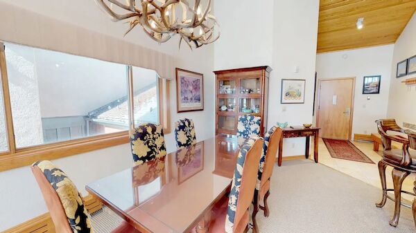 A dining room with floral-upholstered chairs, a glass-top table, an antler chandelier, and a cabinet with glass doors.