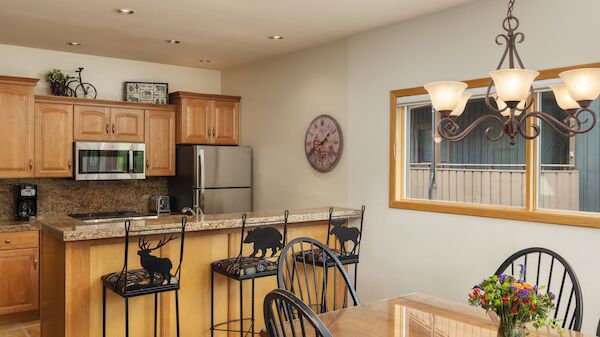A cozy kitchen with wooden cabinets, a dining table, and decorative chairs. A clock and chandelier add charm, along with a vase of flowers.