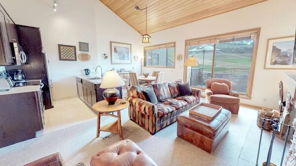 The image shows a cozy living room with a couch, armchairs, kitchen area, and large windows with a scenic view of the outdoors, featuring wooden elements.