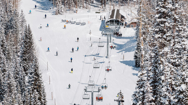 Skiers and snowboarders are on a snowy slope with a chairlift and surrounded by trees. The snowy scene depicts a busy winter day on the mountain.