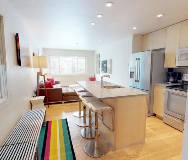 A modern kitchen with an island, bar stools, stainless steel appliances, a colorful rug, and a living area with a couch and a lamp in the background.