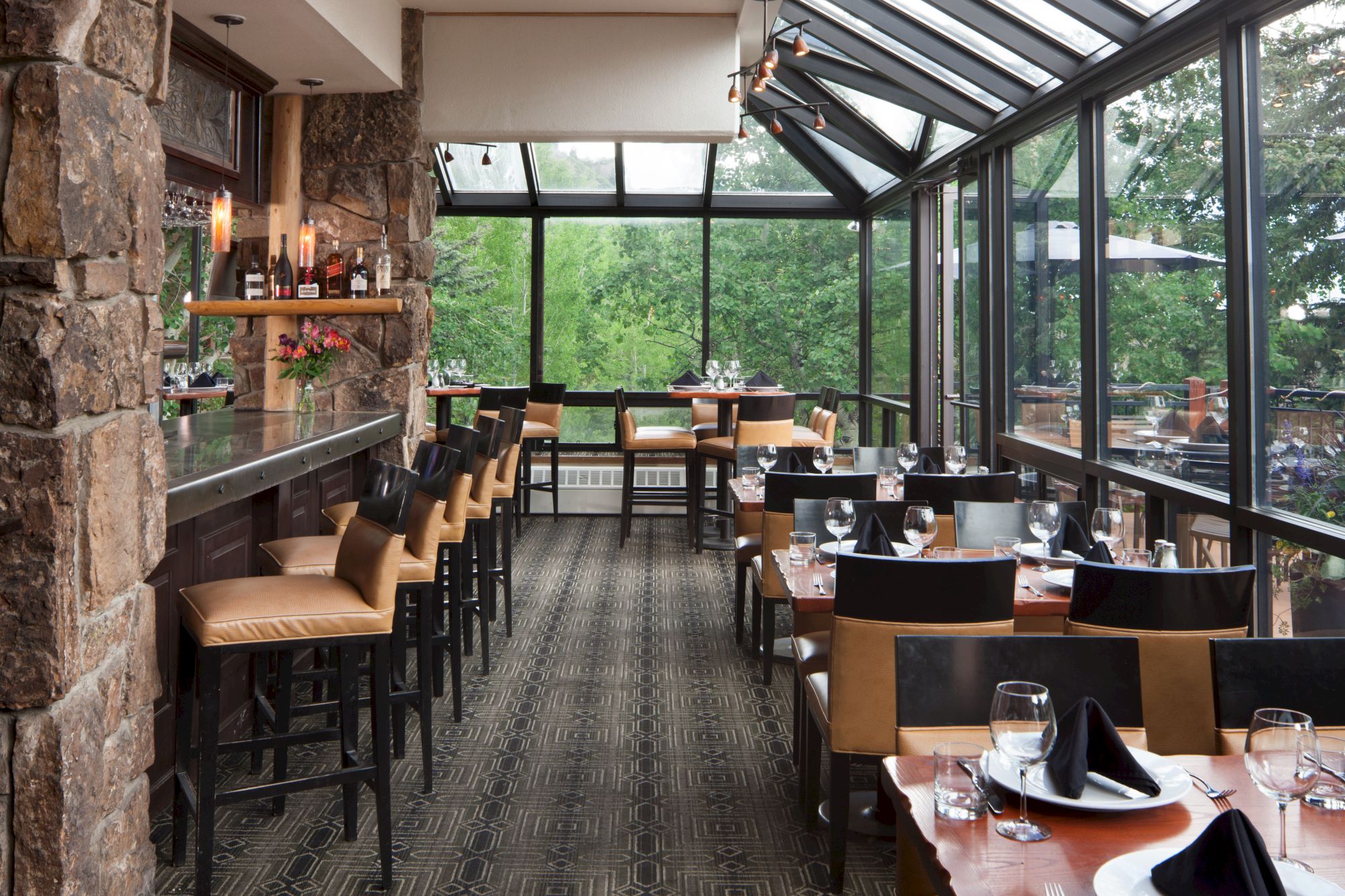 A warmly lit restaurant with a stone wall, bar, and seats on the left, and dining tables with place settings to the right, large windows show greenery outside.