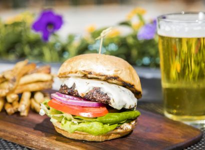 A cheeseburger with lettuce, tomatoes, and onions, served with fries on a board, with a glass of beer in the background.