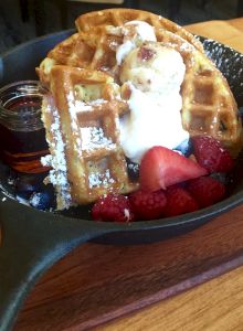 A skillet with waffles, a dollop of whipped cream or ice cream, syrup on the side, powdered sugar, and fresh strawberries and raspberries.