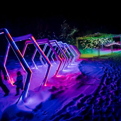 A person and child walk through a snow-covered path adorned with colorful illuminated geometric arches, leading to a lit area ahead.
