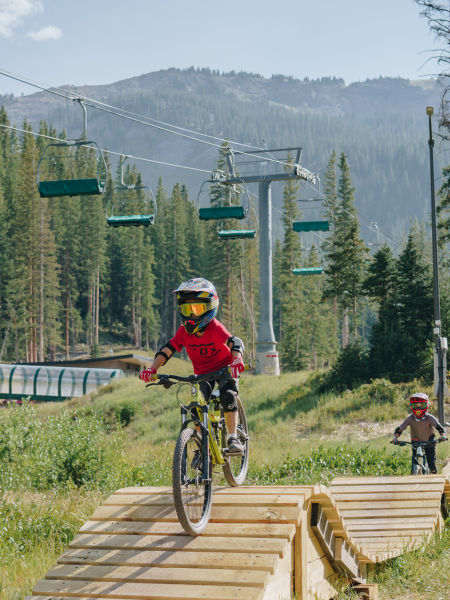 The image shows a group of children riding mountain bikes on a wooden trail in a beautiful forested area with ski lifts and mountains in the backdrop.