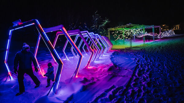 A person and a child are walking through a tunnel made of illuminated hexagonal frames in a snowy, colorful nighttime setting.