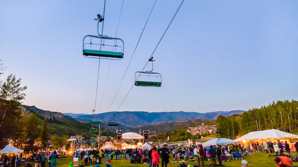 A large outdoor gathering with people seated and standing on a grassy field, surrounded by tents, set against a mountainous backdrop with ski lifts overhead.