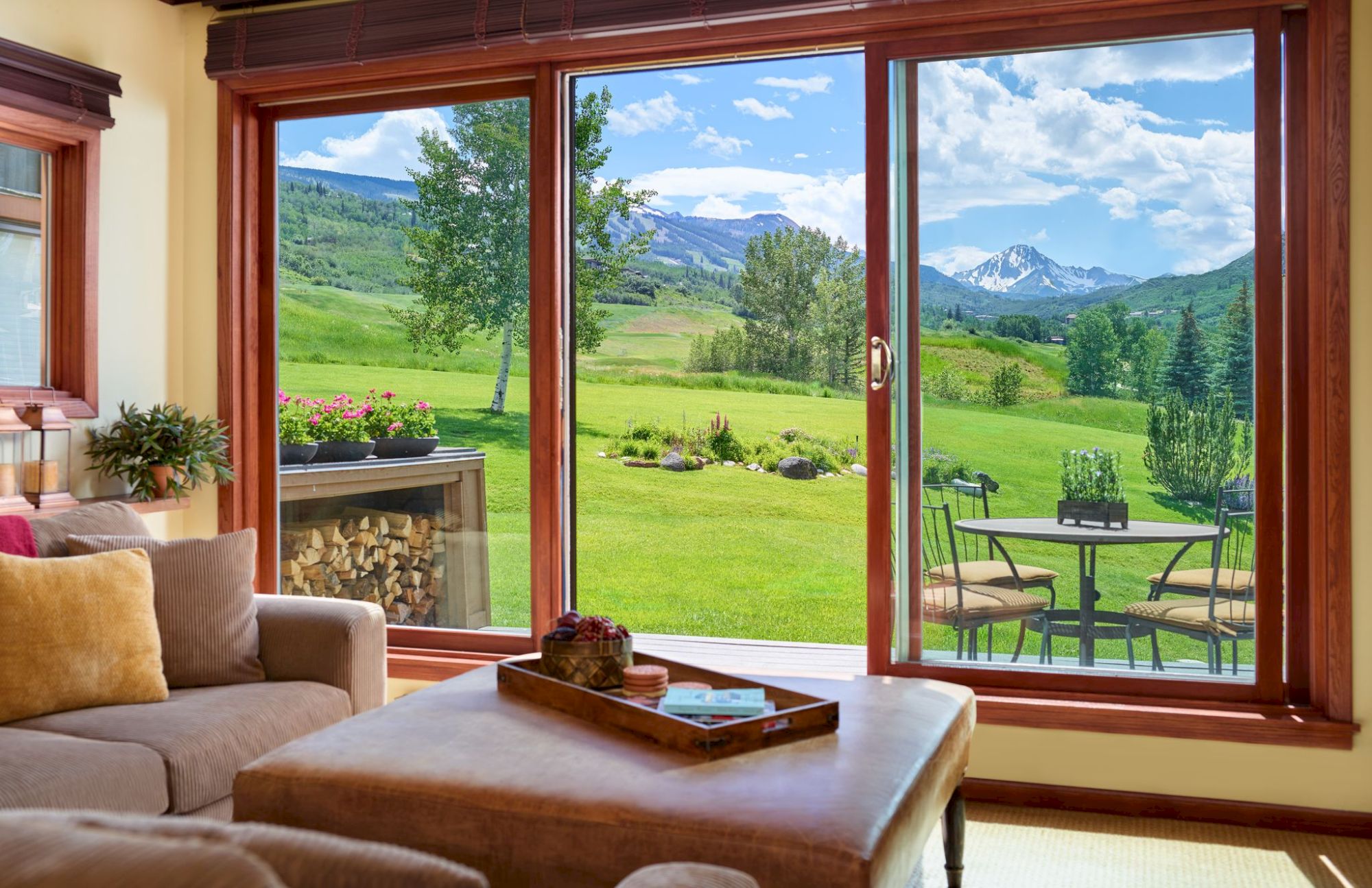 A cozy living room with a view of a lush green lawn, a patio set outside, and mountains in the background through large sliding glass doors.