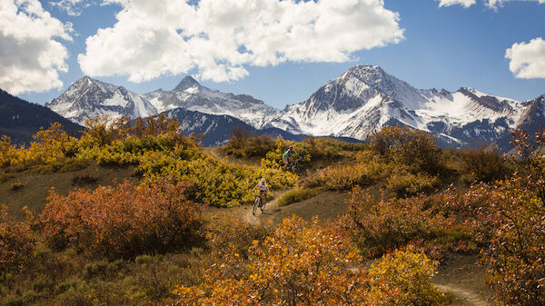 The image features stunning mountains covered in snow, surrounded by colorful autumn foliage, under a bright, cloudy sky.
