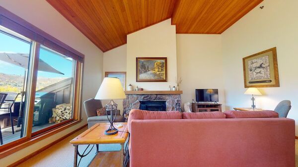 The image shows a cozy living room with wooden ceilings, a fireplace, soft furnishings, and a view through large windows.