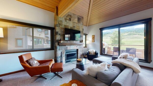 A cozy living room with a stone fireplace, TV, large windows, and modern furniture including a leather chair and a plush sofa.