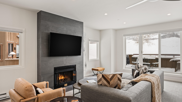 A modern living room with a fireplace, TV, gray sofa, and armchairs, featuring large windows with snow-covered outdoor view.